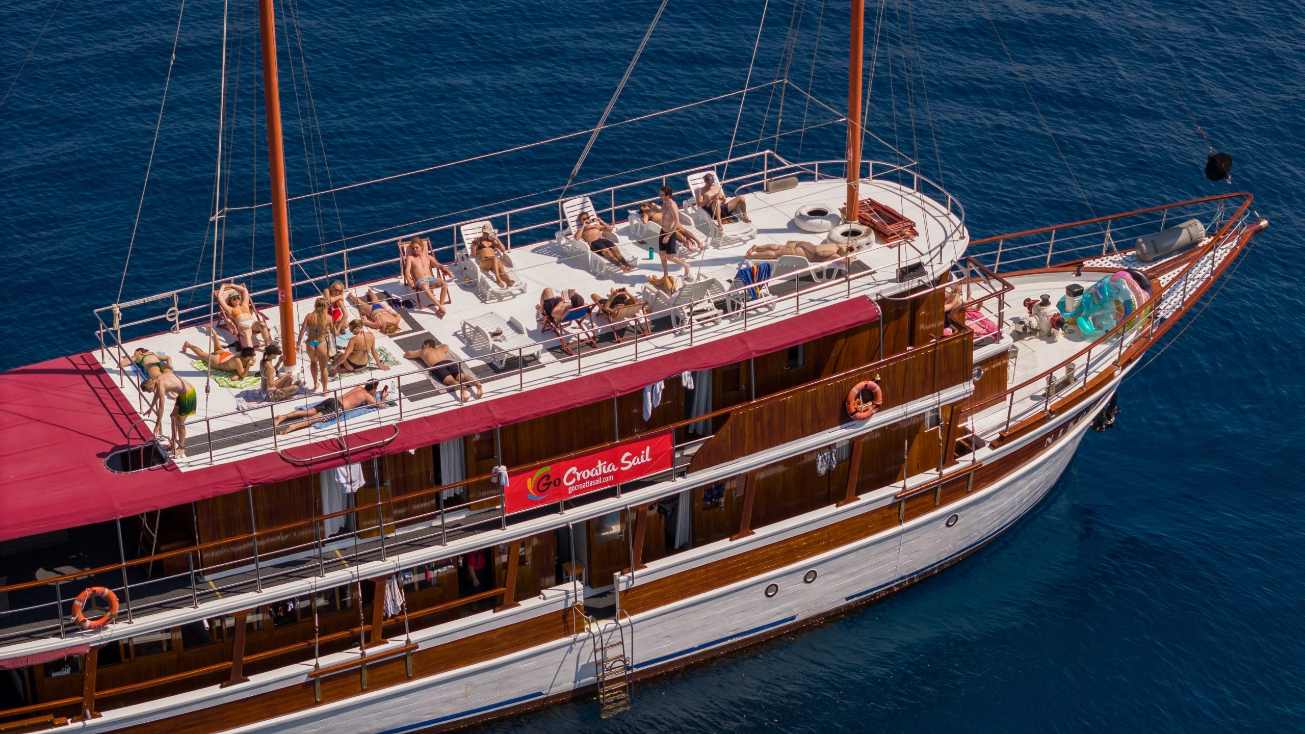An areal close-up shot of a Classic Plus boat with a red Go Croatia Sail banner on a summer day with people sunbathing on the top deck